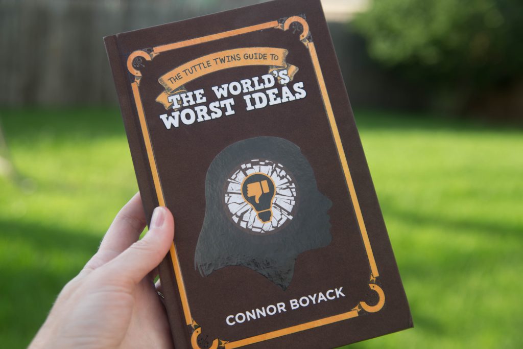 A hand holding a book titled The Tuttle Twins Guide to the World's Worst Ideas by Connor Boyack 